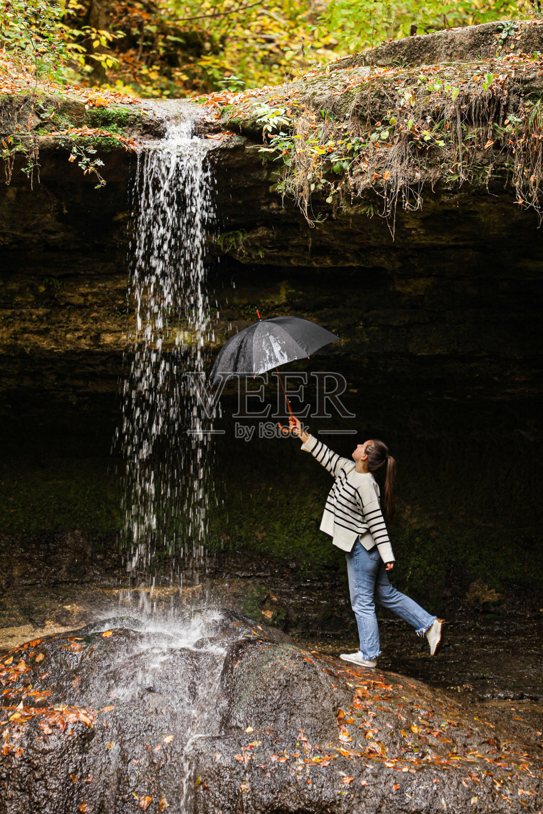 一个徒步旅行的女孩站在一个令人惊叹的热带瀑布上，一把黑色的伞站在瀑布的雨中，孤立在瀑布流过的洞穴的背景上。照片摄影图片