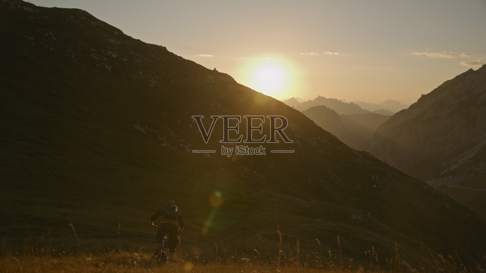 男子骑自行车的后视图在草地上对雄伟的山脉在明亮的日落照片摄影图片