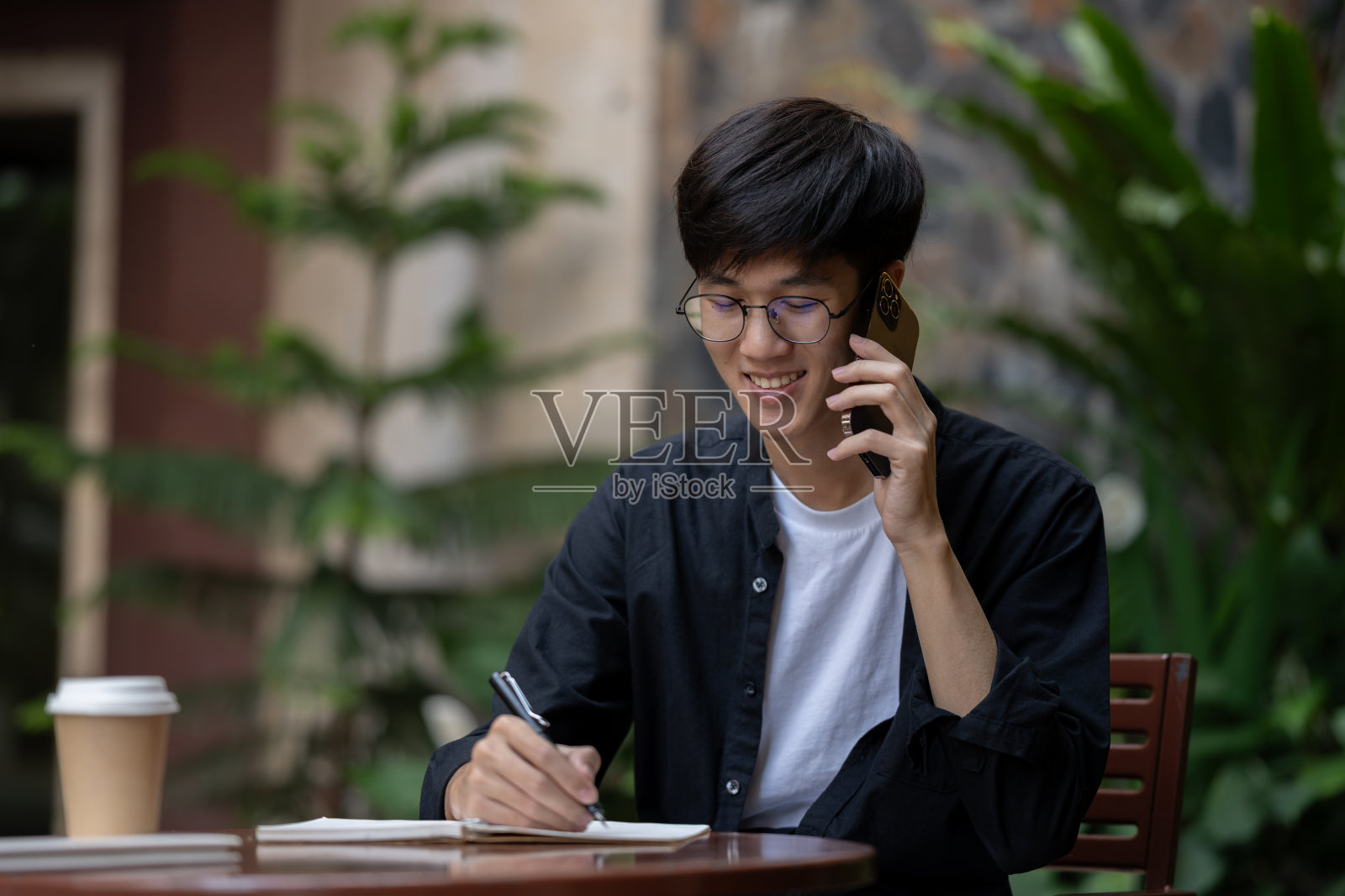 一位快乐的亚洲男子在户外与人通电话时正在记笔记。照片摄影图片