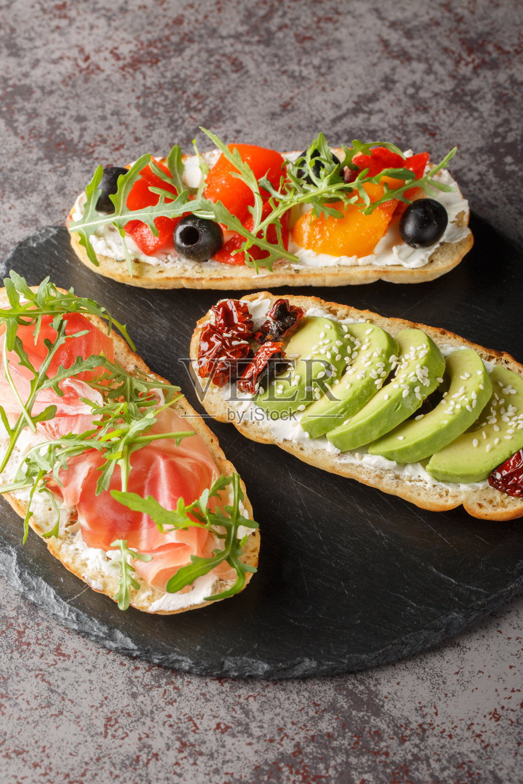 新鲜的开放式三明治，配上芝麻菜、火腿、晒干的西红柿、鳄梨、奶油奶酪、橄榄和烤辣椒。垂直照片摄影图片