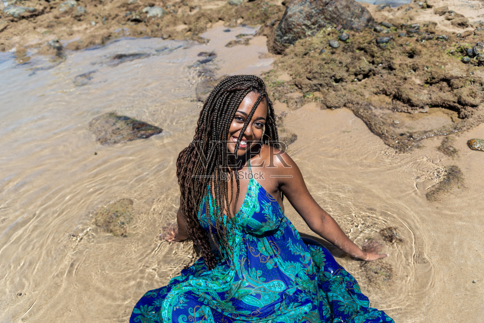 一个美丽的女人的画像，穿着蓝色的连衣裙，扎着辫子，坐在沙滩上摆姿势拍照。在海滩上晒日光浴。照片摄影图片