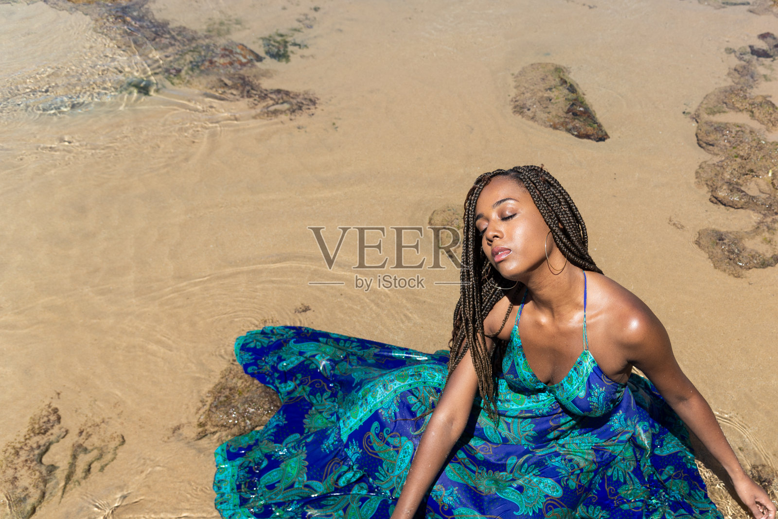 美丽的女人用辫子的头发坐在水里在海滩上晒日光浴的蓝色长裙。照片摄影图片