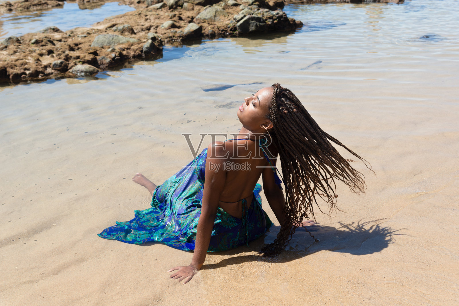 美丽的女人穿着蓝色的裙子坐在沙滩上，她的背部移动着她的辫子。照片摄影图片