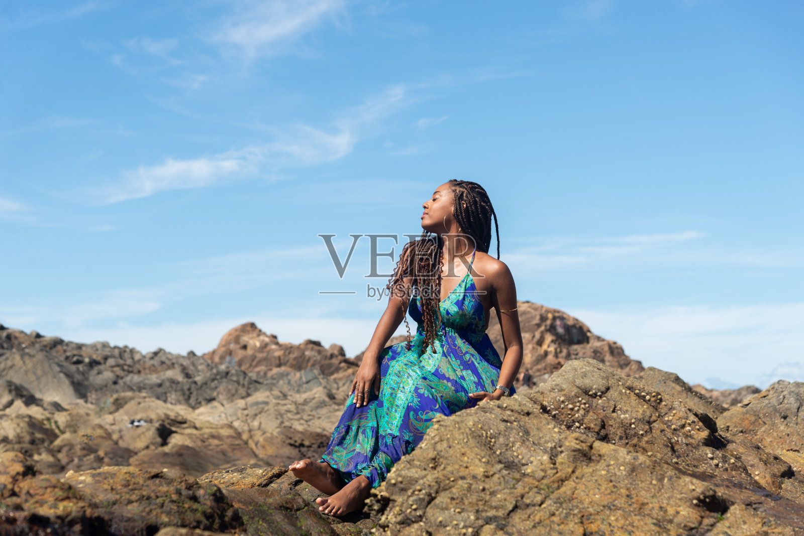 美丽的女人，扎着辫子，穿着蓝色的衣服，坐在海滩的岩石上晒日光浴，摆姿势拍照照片摄影图片