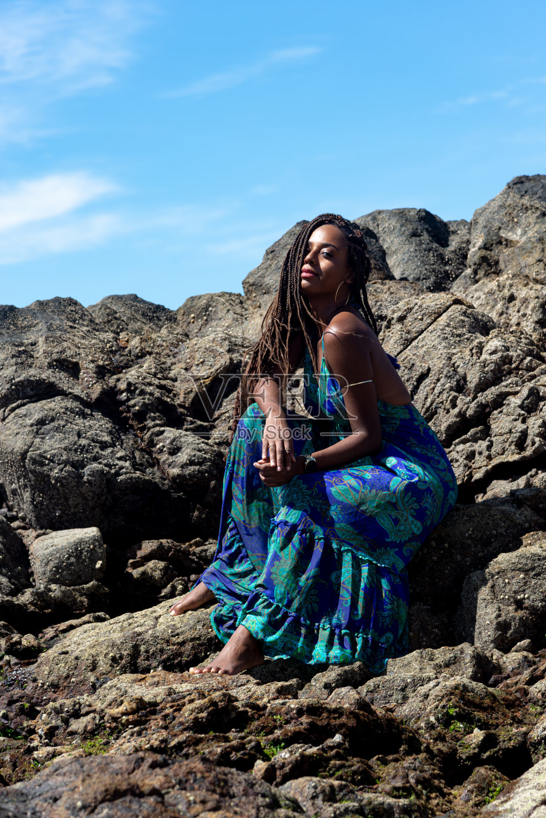 一个漂亮的女人，穿着蓝色的衣服，扎着辫子，坐在海滩的黑色岩石上晒日光浴照片摄影图片