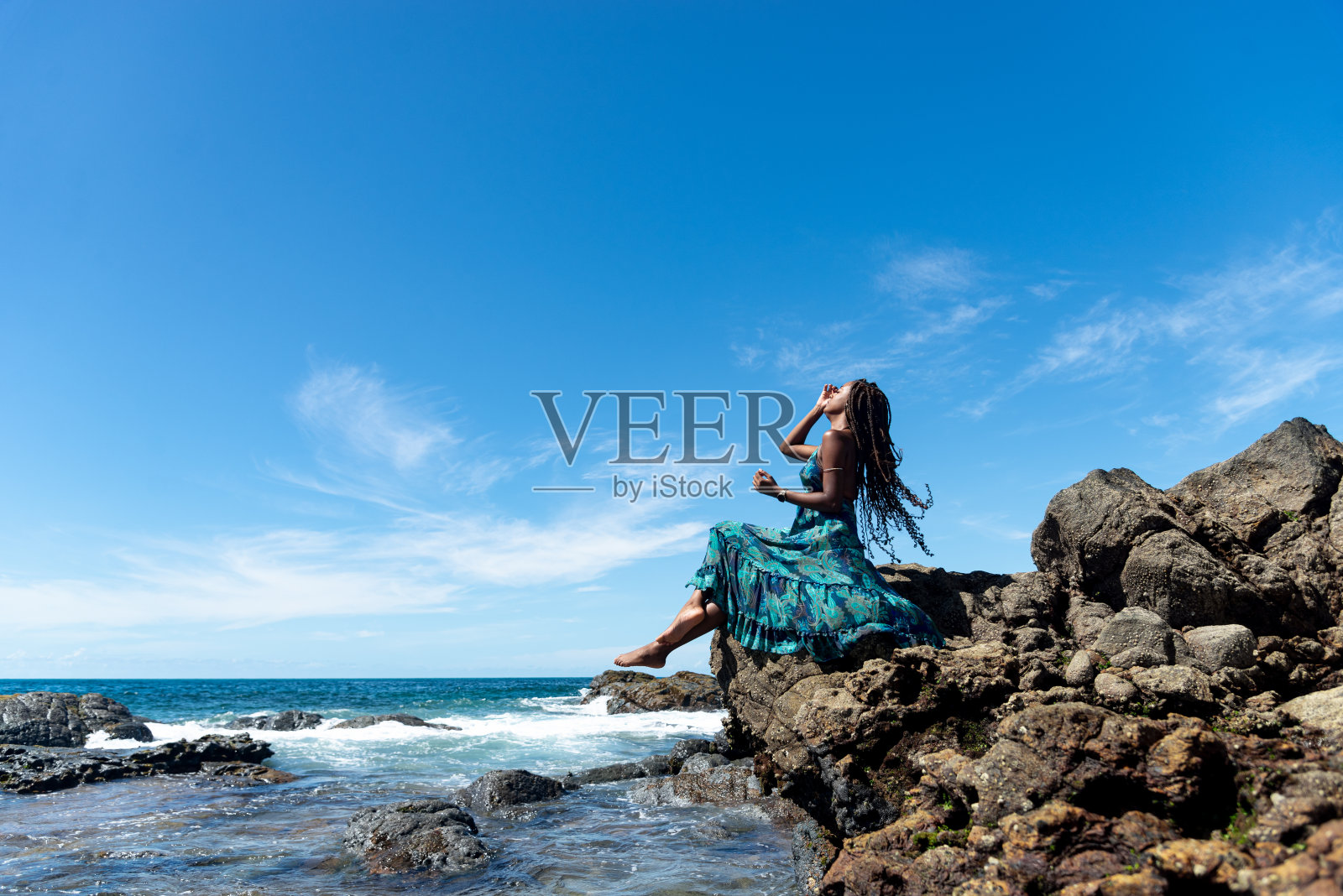 穿着蓝色衣服、扎着辫子的漂亮女人坐在海滩岩石边晒太阳照片摄影图片