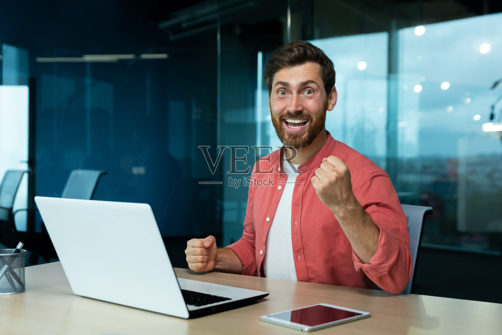 办公室里穿着红衬衫的成功成熟商人的肖像，在工作中庆祝胜利和成功的成果，看着相机，举起手照片摄影图片