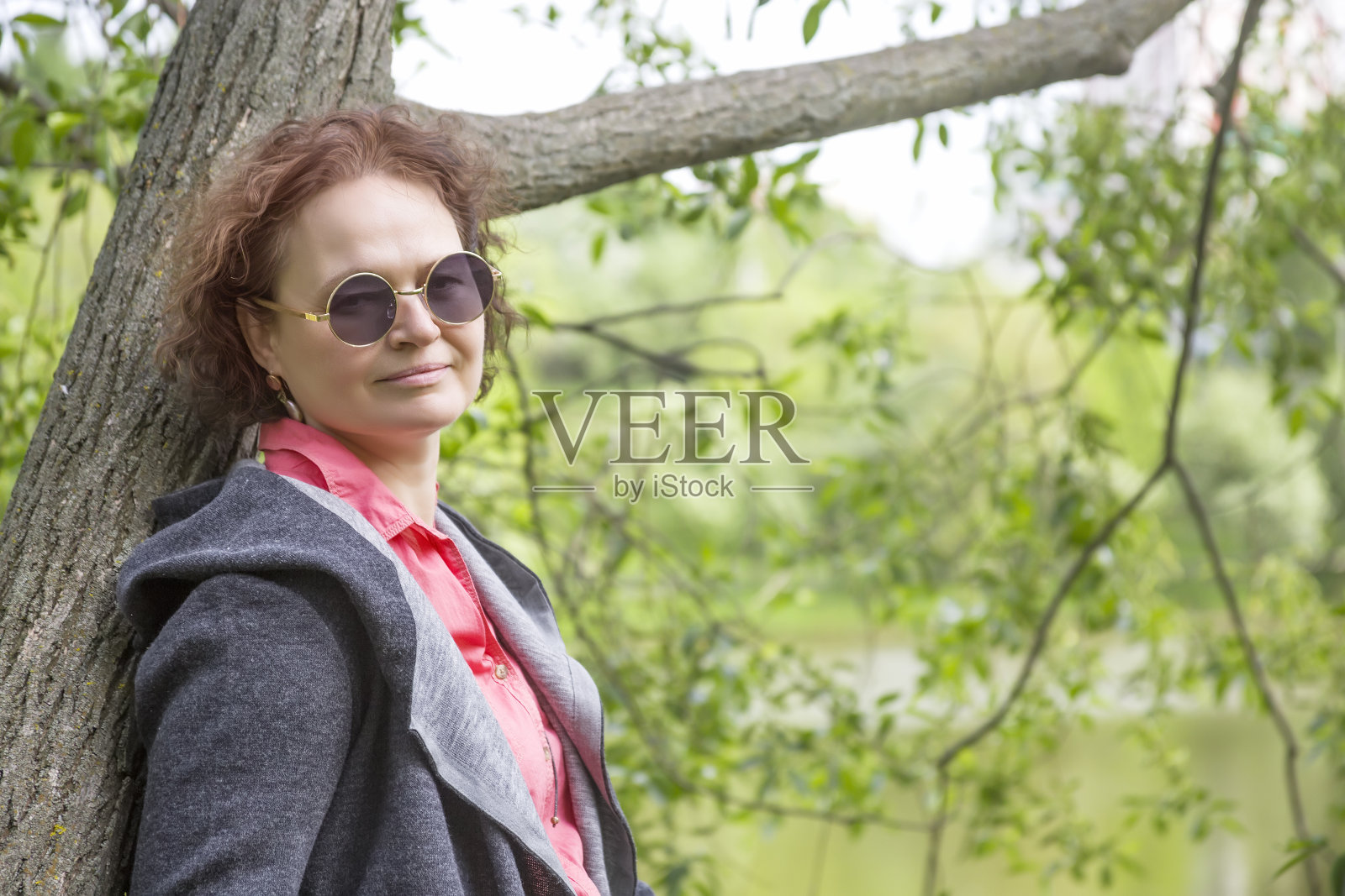 一个戴着墨镜的成熟女人靠在一棵树上看着镜头。背景-春天的树叶照片摄影图片