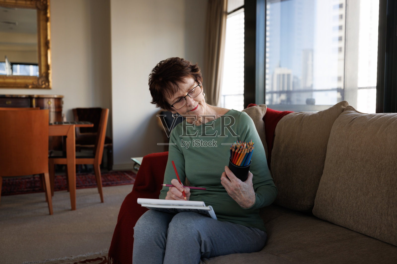 拿着记事本的高级妇女坐在家里的沙发上看着她的彩色铅笔。照片摄影图片