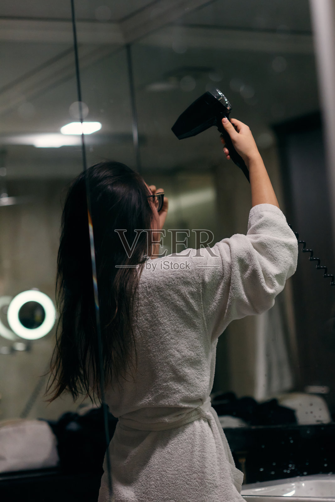 沐浴后，一个快乐的女人戴着耳机，在浴室里用吹风机吹干头发，这是一个无忧无虑、充满活力的自我护理和放松时刻照片摄影图片