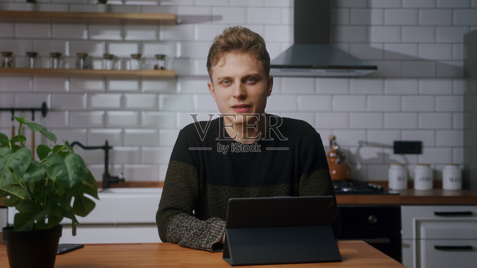 年轻的金发视频博主带着平板电脑坐在家里的现代厨房里，看着相机，说话，录制视频照片摄影图片