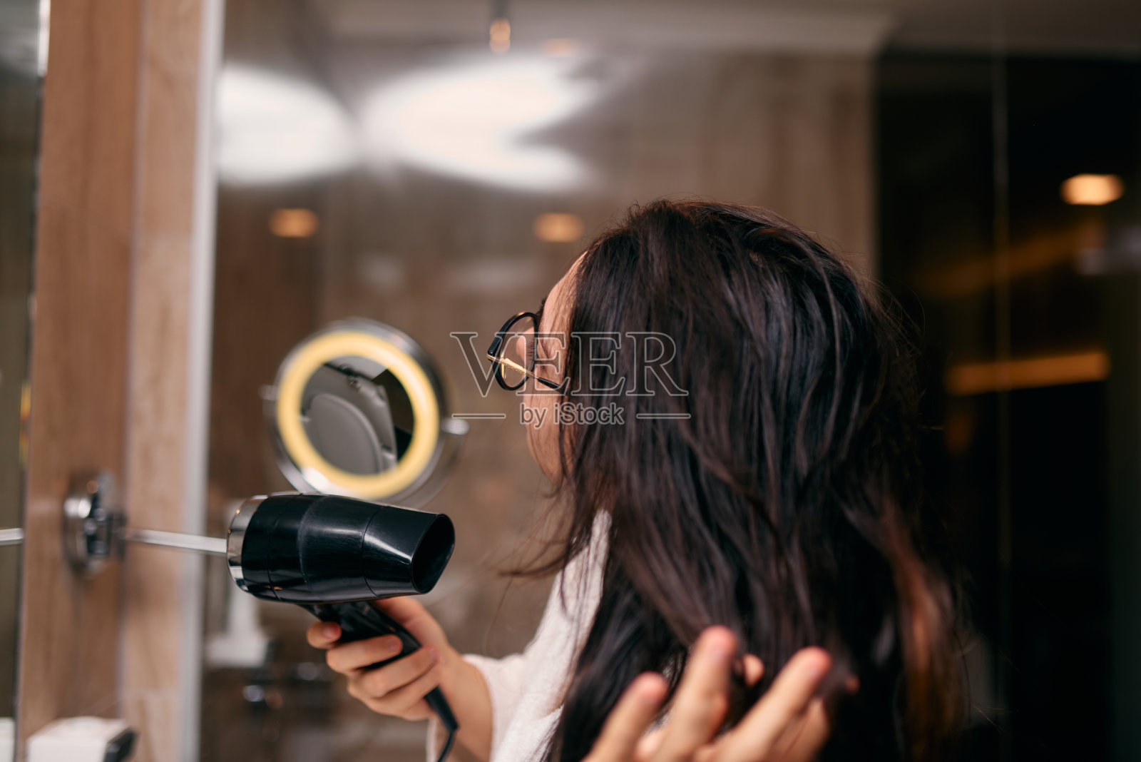 沐浴后，一个快乐的女人戴着耳机，在浴室里用吹风机吹干头发，这是一个无忧无虑、充满活力的自我护理和放松时刻照片摄影图片