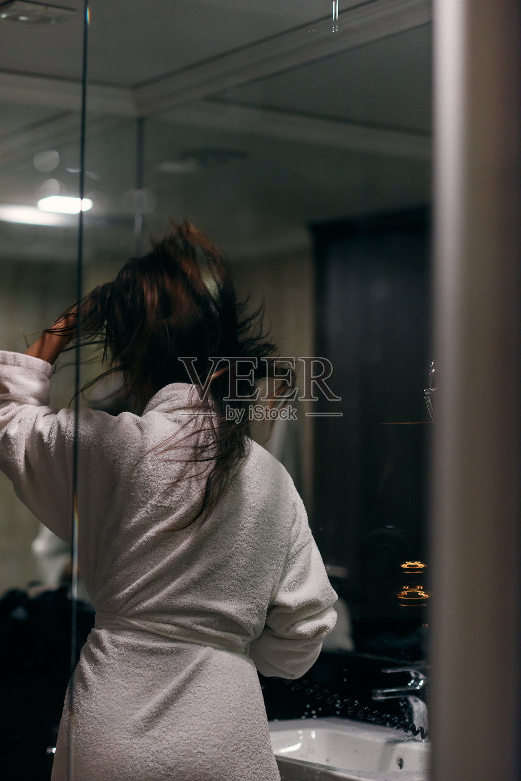 沐浴后，一个快乐的女人戴着耳机，在浴室里用吹风机吹干头发，这是一个无忧无虑、充满活力的自我护理和放松时刻照片摄影图片