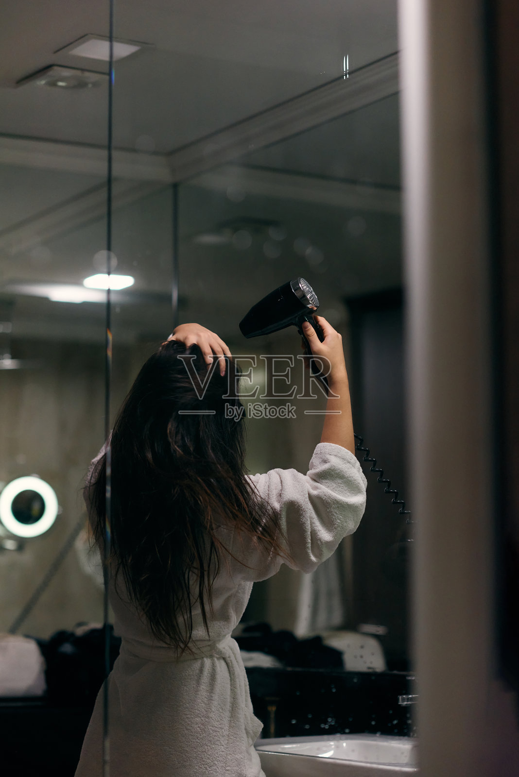 沐浴后，一个快乐的女人戴着耳机，在浴室里用吹风机吹干头发，这是一个无忧无虑、充满活力的自我护理和放松时刻照片摄影图片