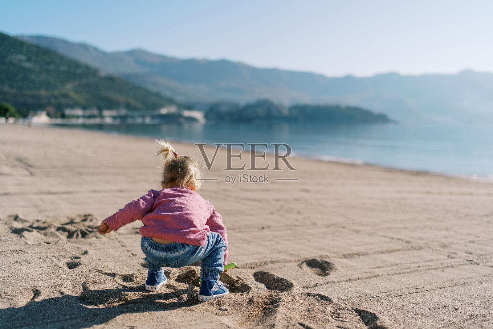 小女孩蹲在沙滩上用铲子挖沙子。后视图照片摄影图片