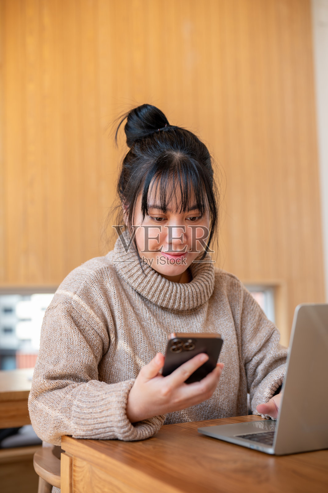 一位乐观的亚洲女性在室内一边用笔记本电脑工作，一边查看智能手机上的信息。照片摄影图片