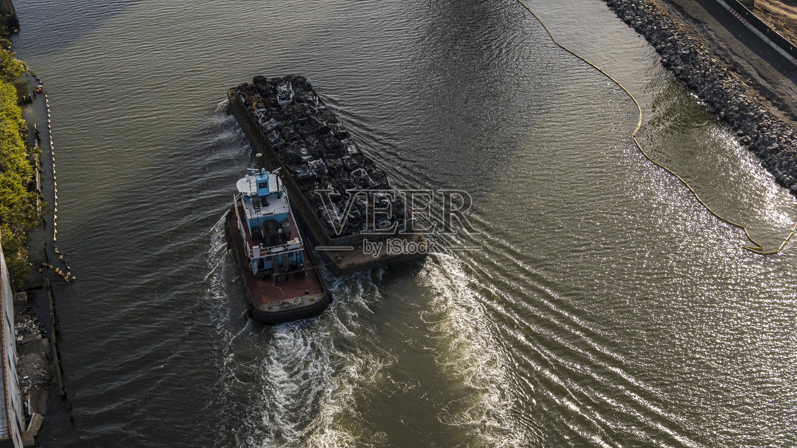 一艘拖船拖着一艘载着废金属的驳船，沿着纽约布鲁克林绿点的纽敦溪行驶。照片摄影图片