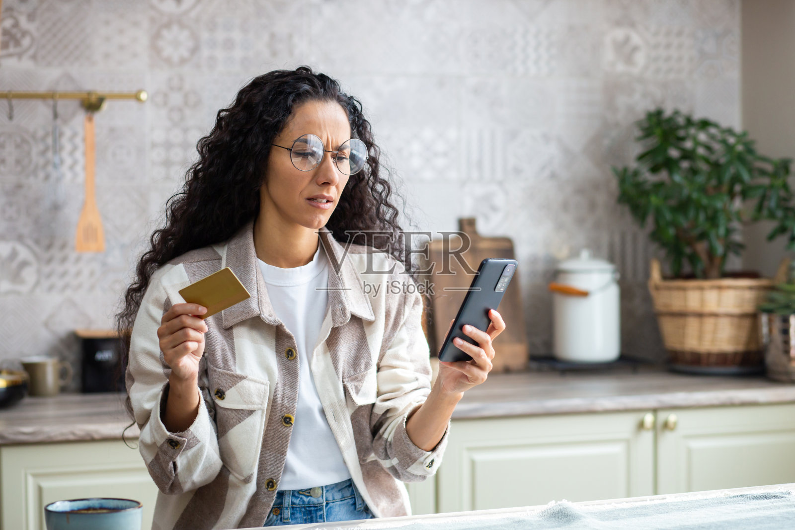 在家中厨房感到沮丧的受骗妇女，银行转账错误，遭遇网络诈骗，伤心的拉丁美洲妇女手持银行信用卡和手机，使用在线购物APP照片摄影图片