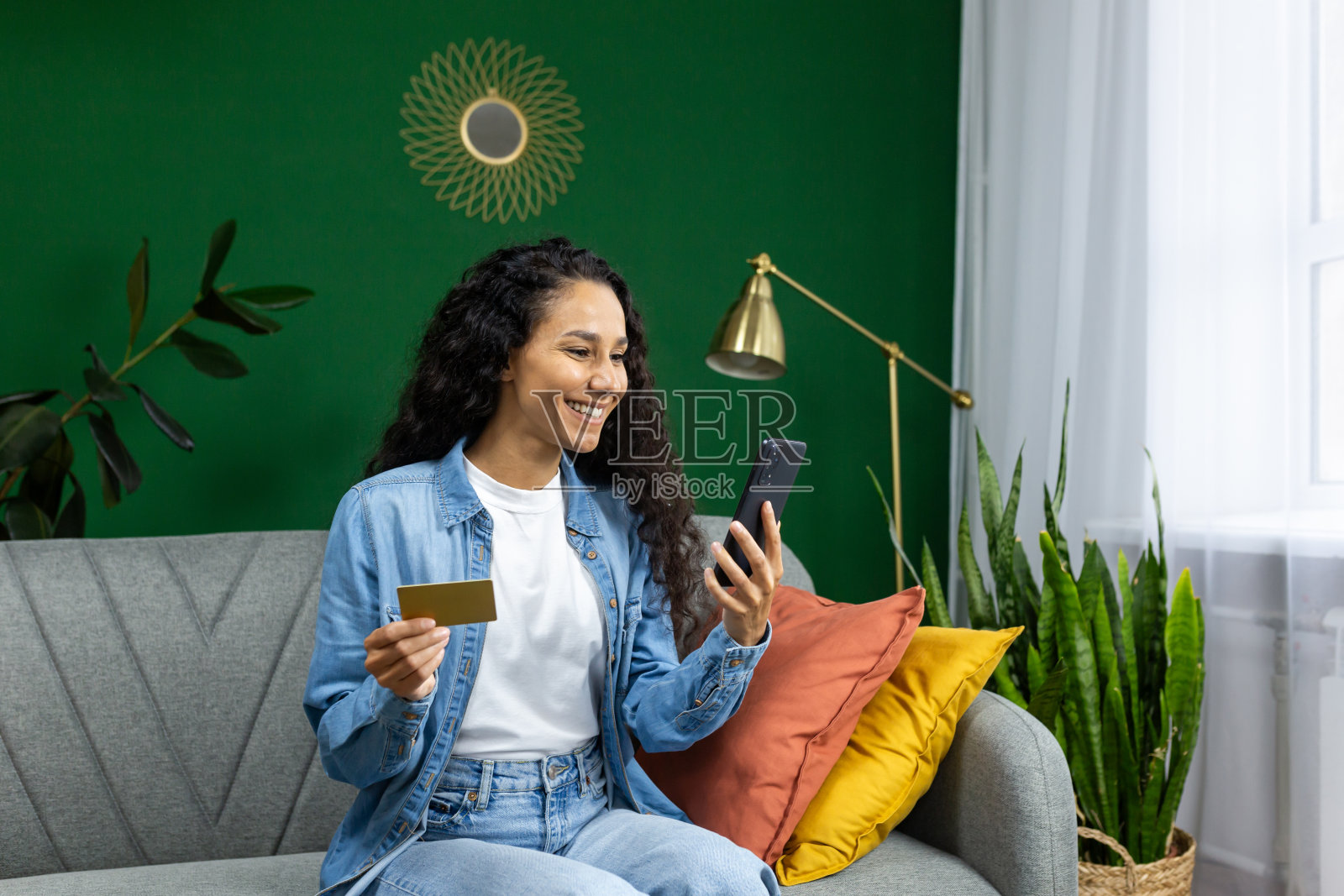 坐在家里的沙发上，手里拿着手机和银行信用卡借记卡的年轻漂亮的快乐女子照片摄影图片