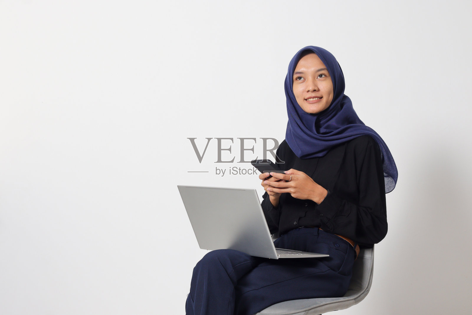 穿着休闲衬衫的亚洲头巾妇女坐在椅子上，一边用笔记本电脑工作，一边用手机。商人的概念。白色背景上的孤立图像照片摄影图片