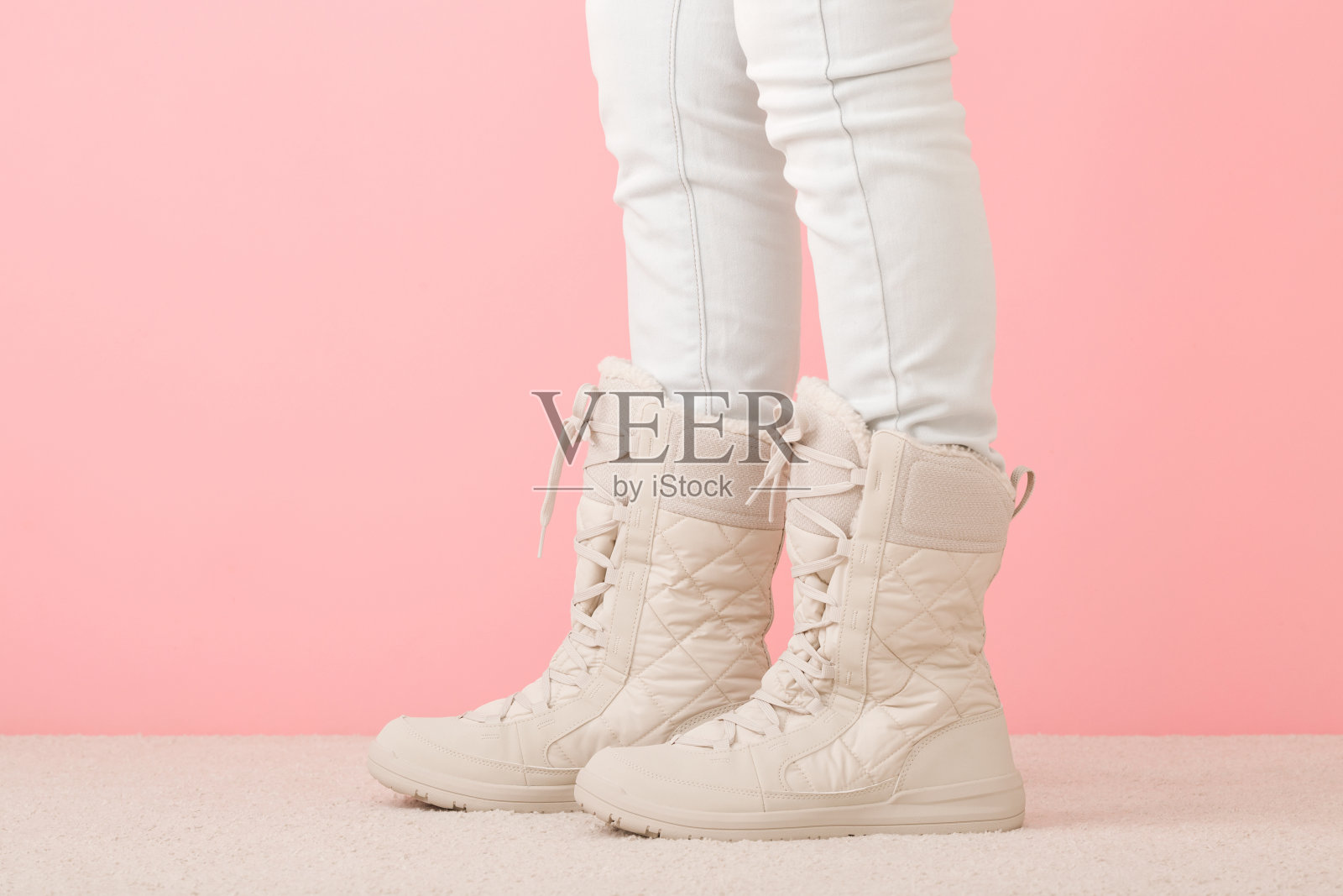 穿着白色牛仔裤和米色暖冬靴的长腿女孩站在粉红色墙壁背景的浅色地毯上摆姿势。柔和的颜色。特写镜头。侧视图。照片摄影图片