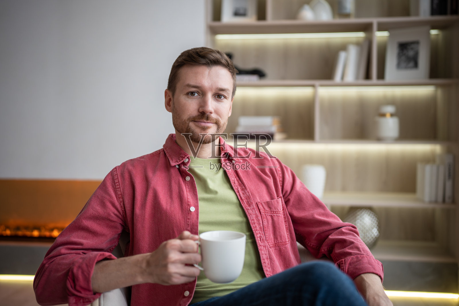快乐友好微笑的男人坐在舒适的公寓里，喝着茶，休息着，看着相机照片摄影图片
