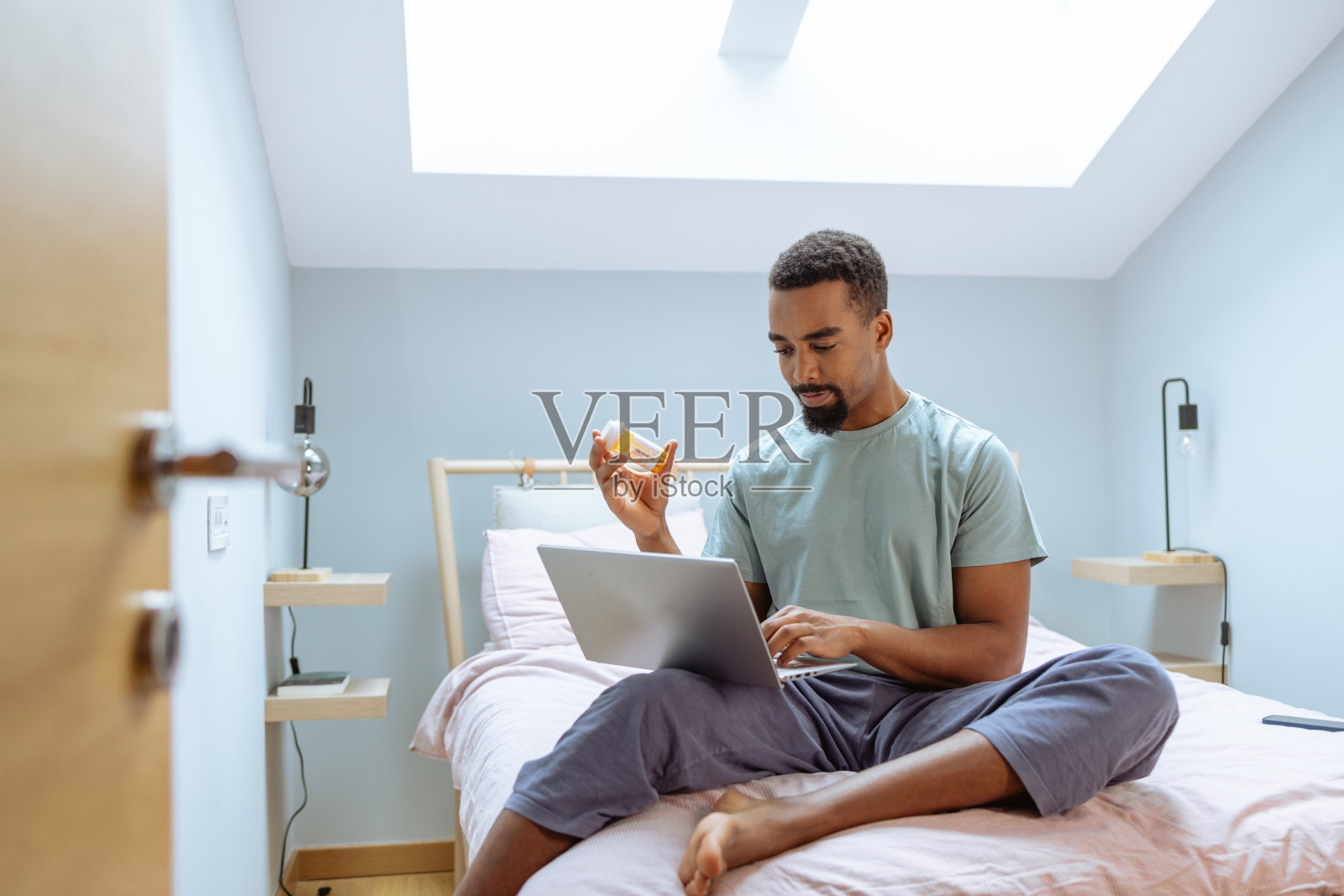 年轻的非裔美国人在卧室里用笔记本电脑，拿着药瓶照片摄影图片