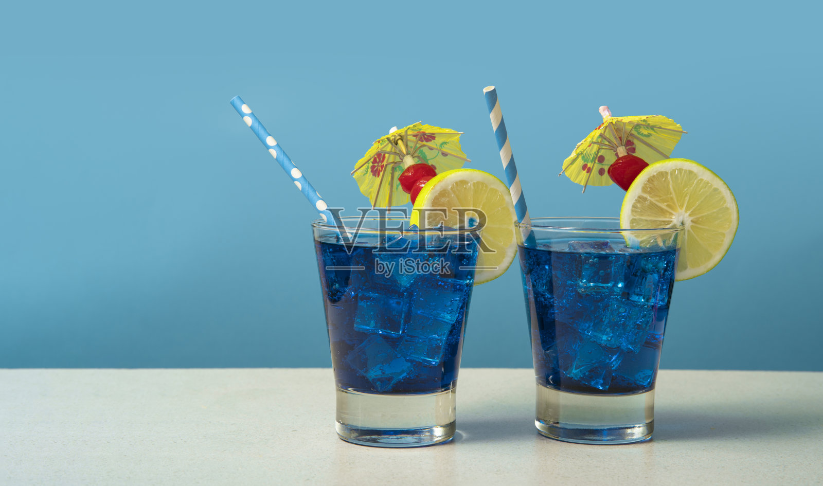 两杯蓝色鸡尾酒，斟满冰块，点缀着柠檬片、樱桃、黄色雨伞和冰糕，放在一张白色的桌子上照片摄影图片