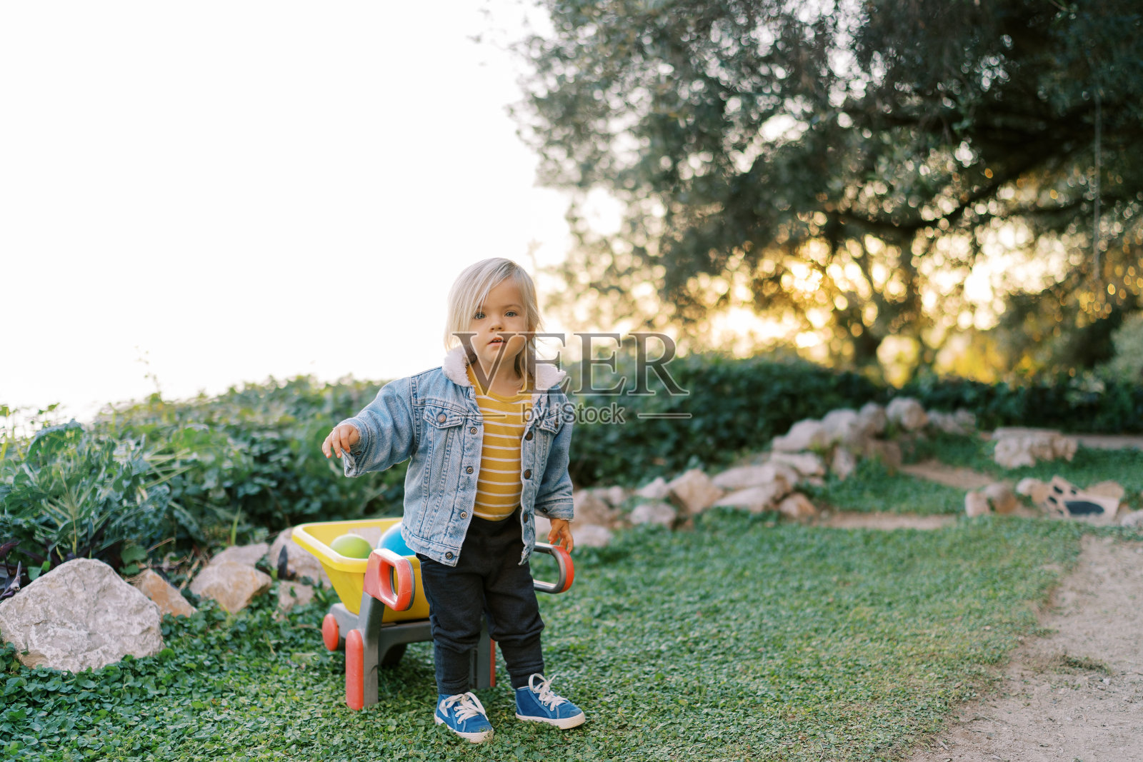 空地上，一个小女孩站在一辆玩具手推车旁边，手推车上放着玩具照片摄影图片