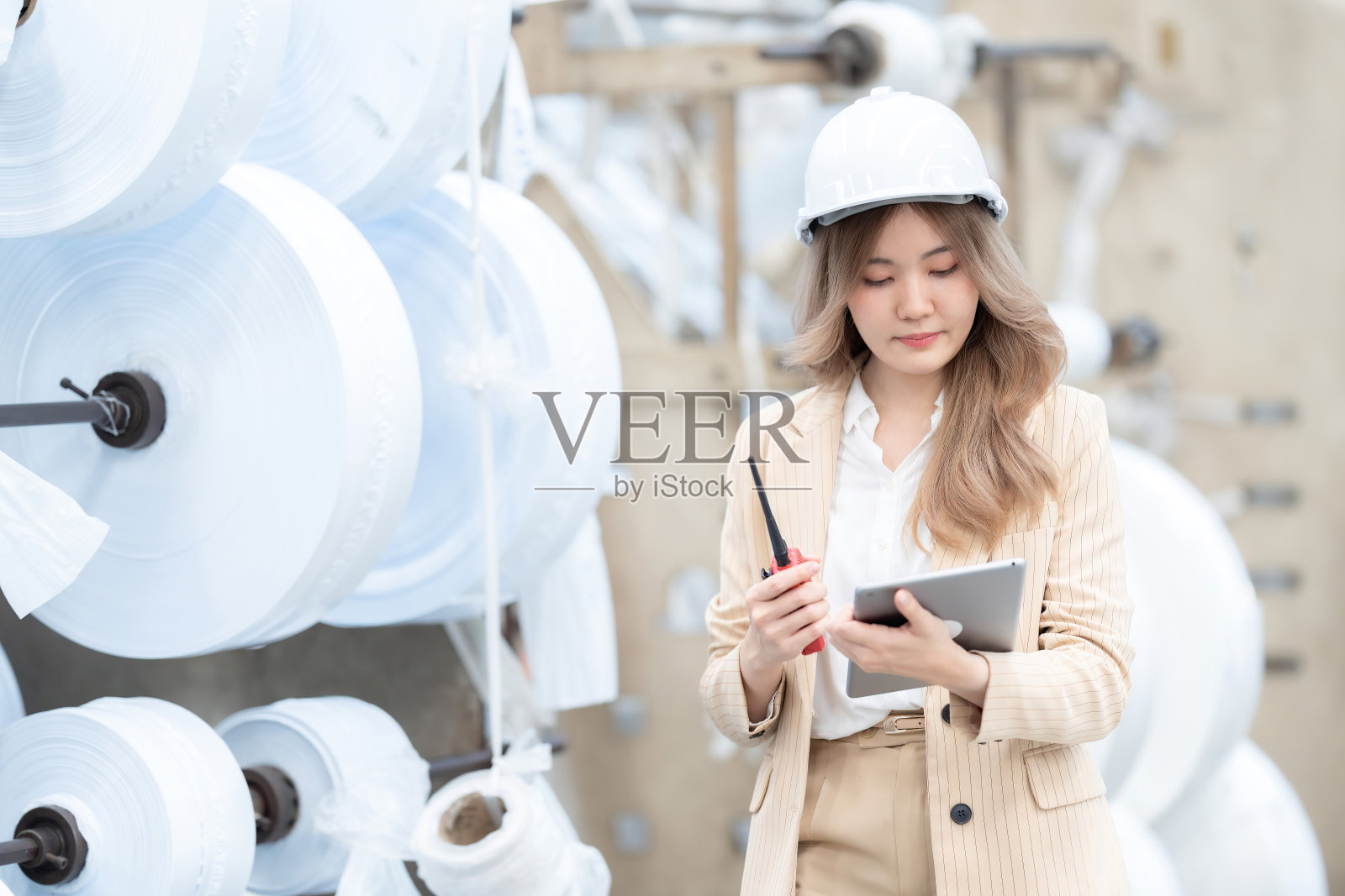 亚洲女性工程师主管使用平板电脑，拿着收音机，戴着安全帽，穿着西装，控制着工业工厂的工作系统。有纸卷作为产品库存。照片摄影图片