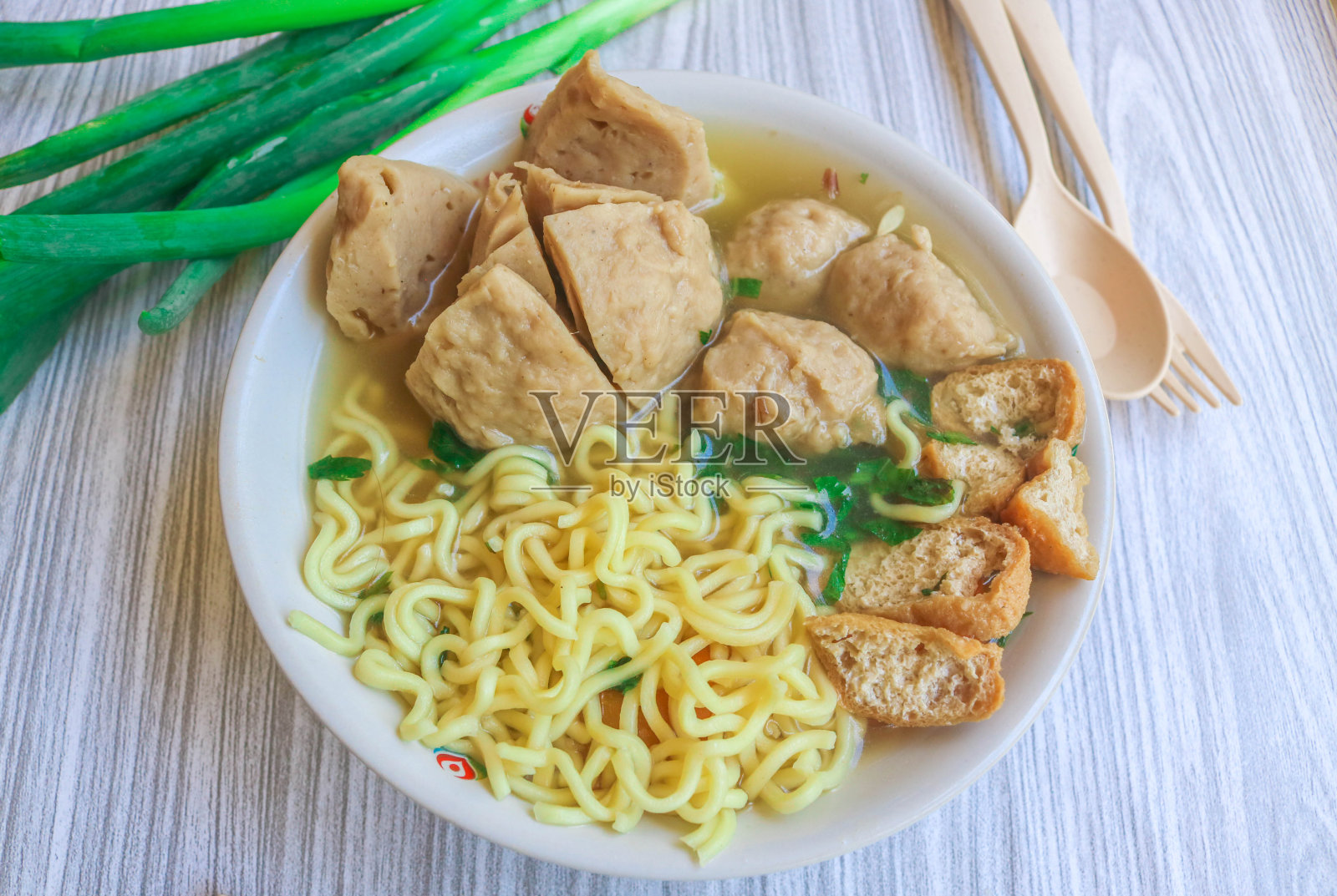 印尼烹饪汤肉丸，里面有面条、豆腐、芹菜和鸡蛋，装在白色的碗里，放在灰色的木桌上。用筷子和勺子在碗里蘸酱的肉丸特写照片摄影图片