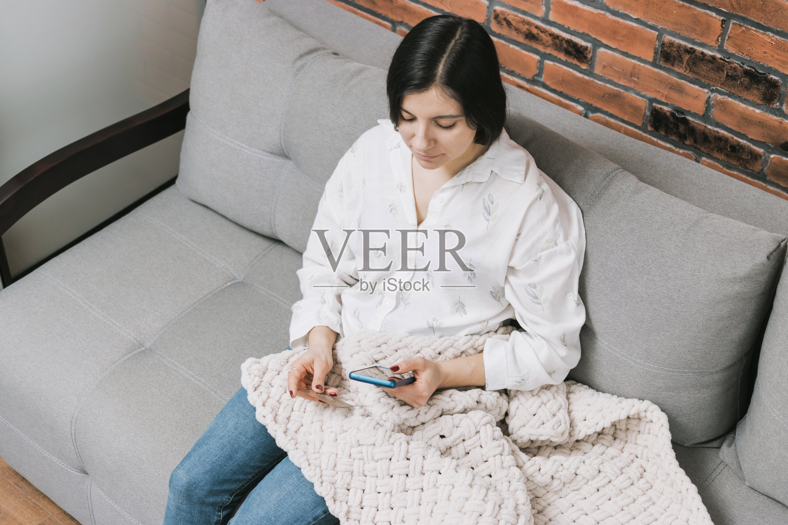 漂亮的年轻女子盖着毯子坐在沙发上，拿着信用卡，用手机、智能手机进行网上购物或网上支付照片摄影图片