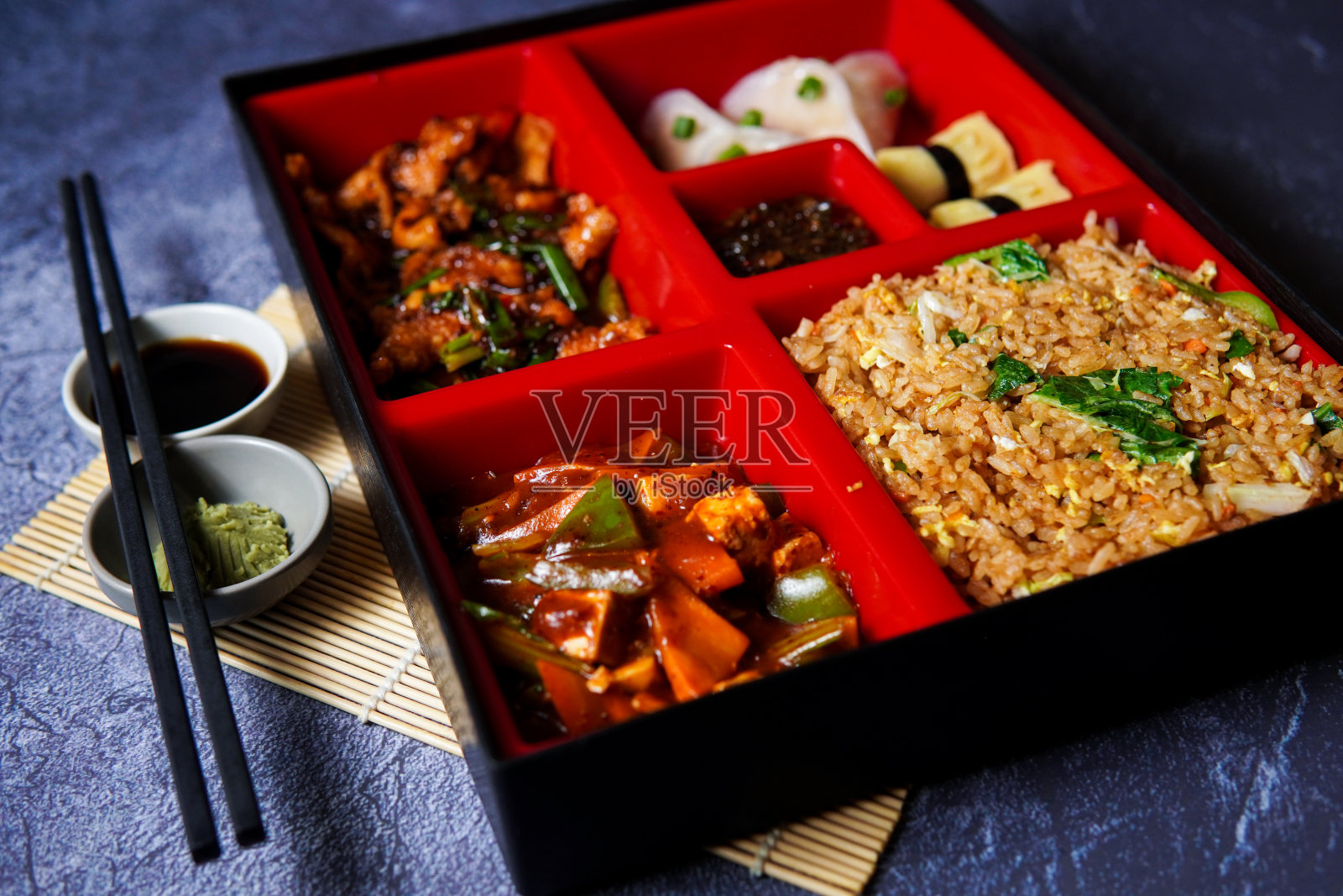 便当盒，用芥末、酱油和筷子做背景。内含炒饭、鸡肉、牛肉、寿司、饺子、辣椒酱、蛋黄酱、美食摄影、社交媒体贴照片摄影图片