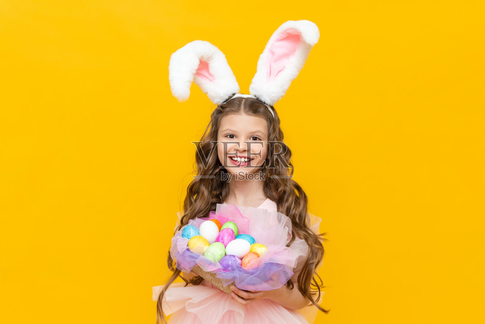 为孩子们准备的节日复活节。一个漂亮的卷发少女拿着一篮子彩色彩蛋准备过节。一个孩子穿着兔子服装，在黄色孤立的背景上长着耳朵照片摄影图片