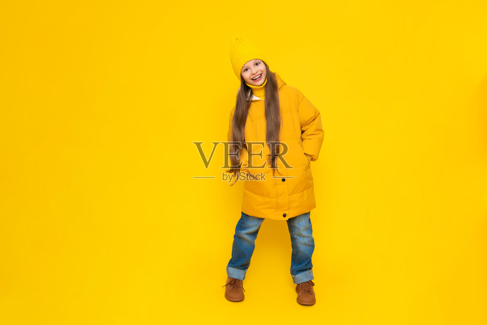 一个小女孩穿着暖和的夹克，戴着帽子，高高兴兴地迎接秋天的来临。美丽的年轻快乐的女孩在一个黄色孤立的背景在工作室。照片摄影图片