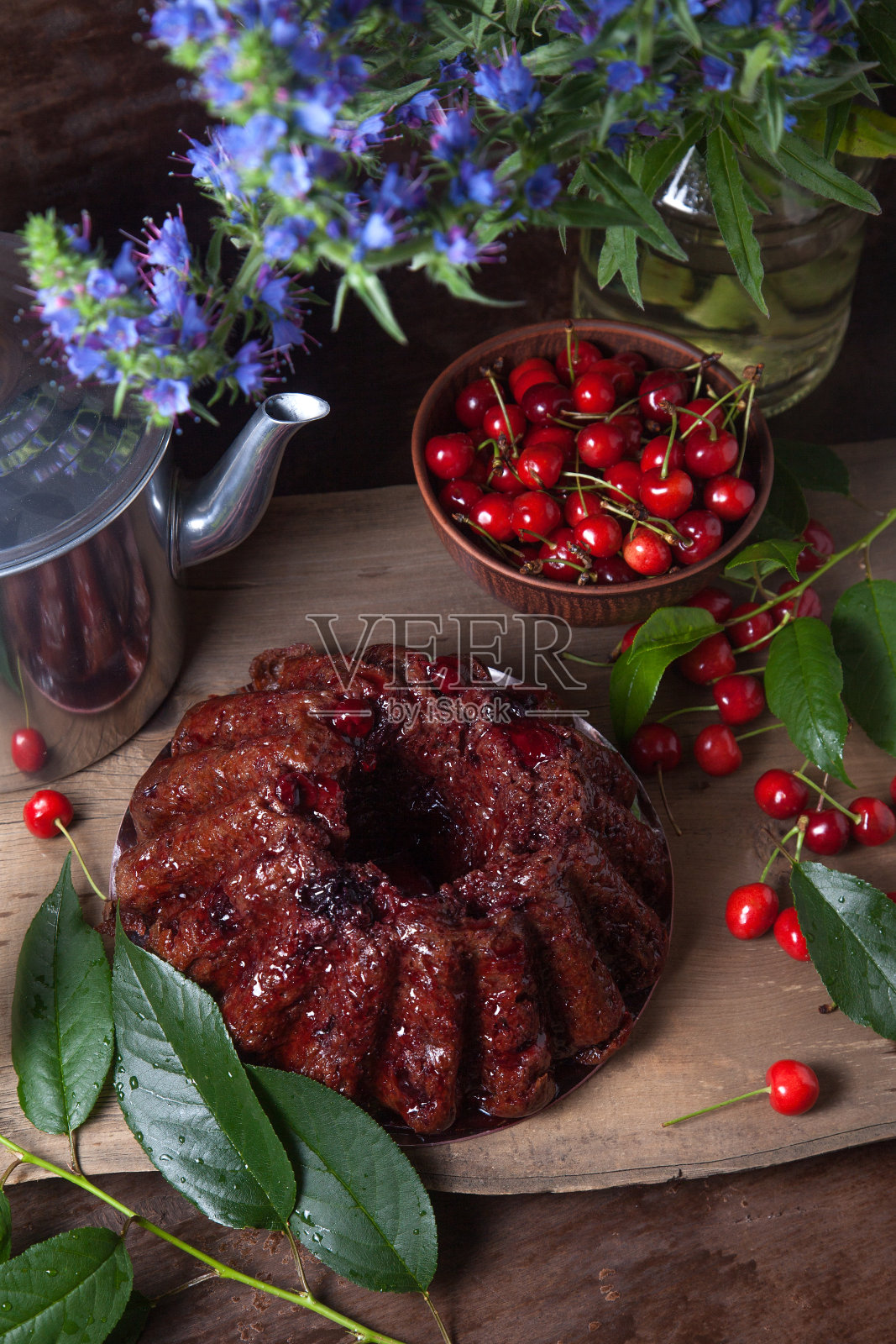 美味的巧克力蛋糕与甜樱桃的浆果，复古水壶和一束蓝色的花在深色木制背景。照片摄影图片