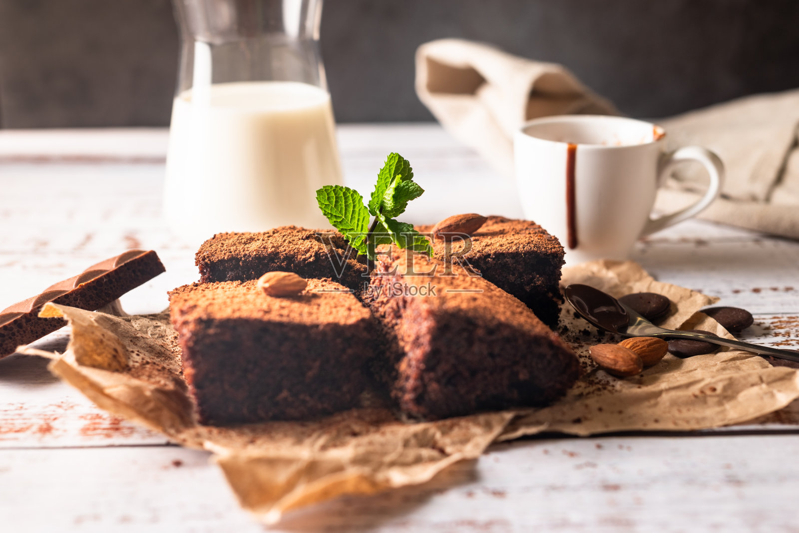 许多新鲜的巧克力布朗尼，撒上可可，放在烘焙纸上，白色的桌子上放着一杯牛奶。自制美味巧克力派照片摄影图片