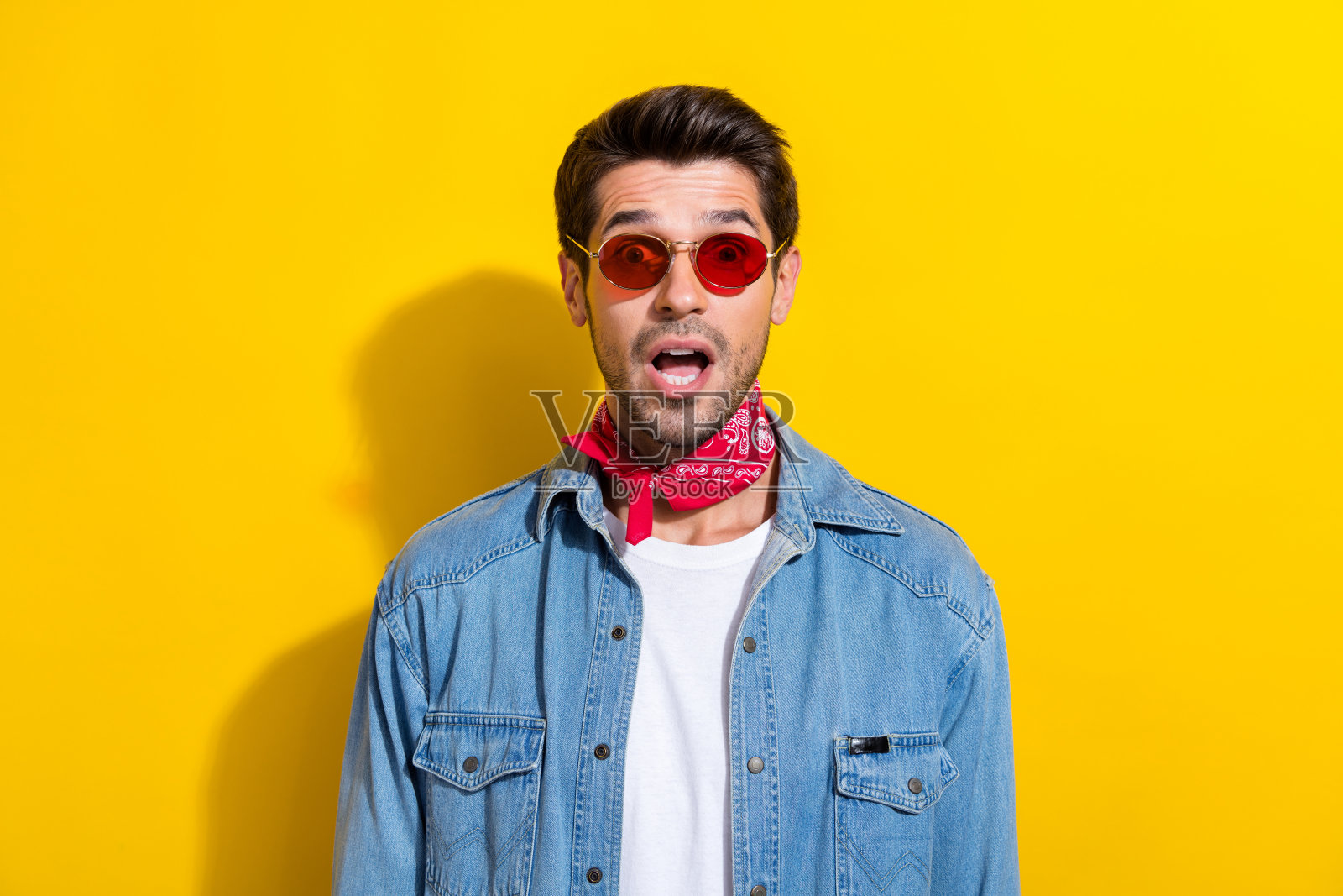 惊讶的男子穿着牛仔裤夹克，戴着眼镜，惊讶地盯着命题张开嘴孤立在黄色背景照片摄影图片