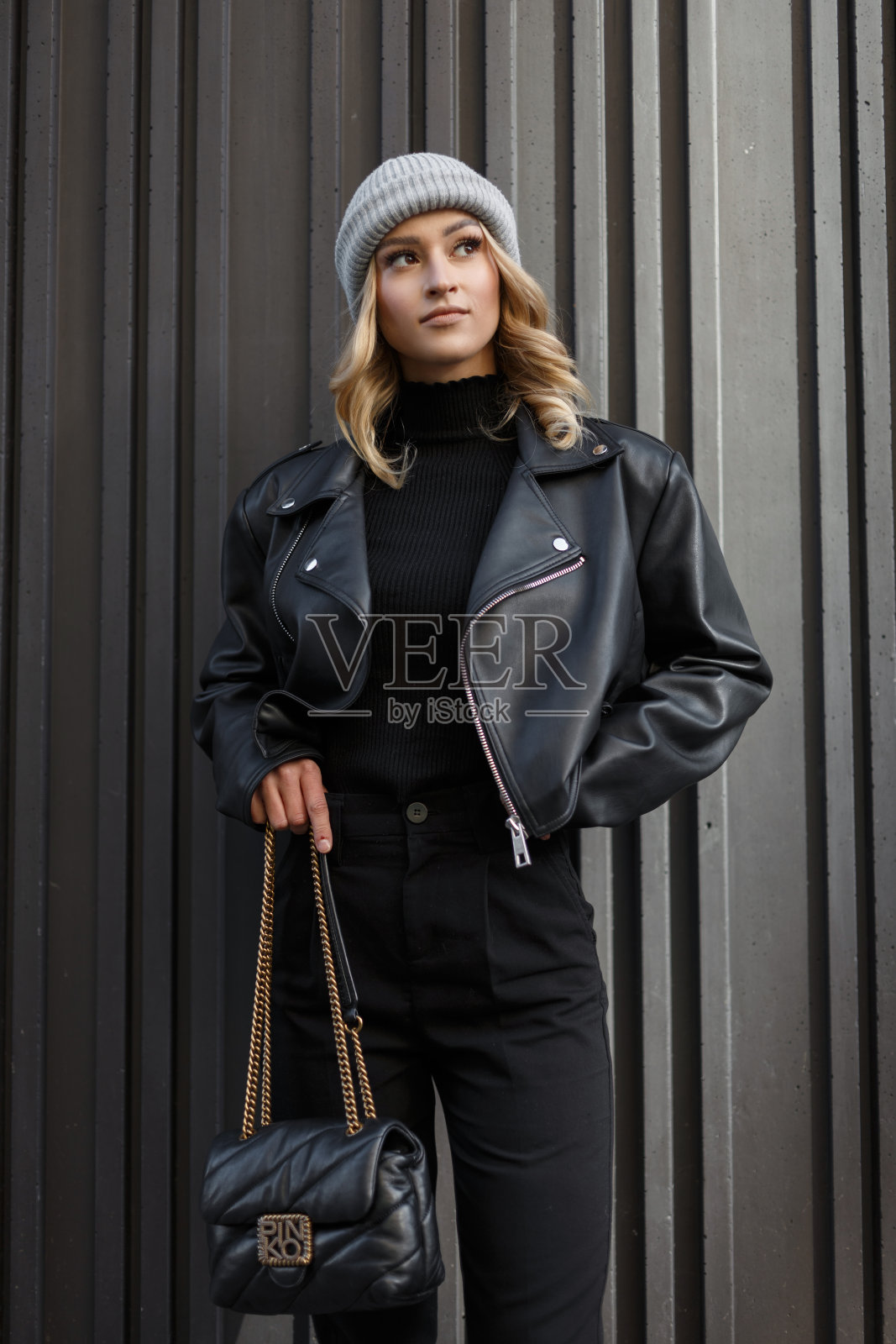 时尚美丽的年轻女子模特，穿着时尚的黑色衣服，皮夹克，毛衣和裤子，戴着针织帽，拿着时尚的黑色手提包，站在城市的黑色墙壁旁。城市照片摄影图片