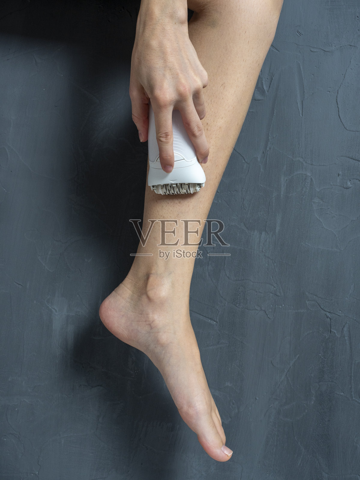 一位女士在灰色纹理背景上用电动脱毛器刮腿毛。美丽和自我护理的概念照片摄影图片