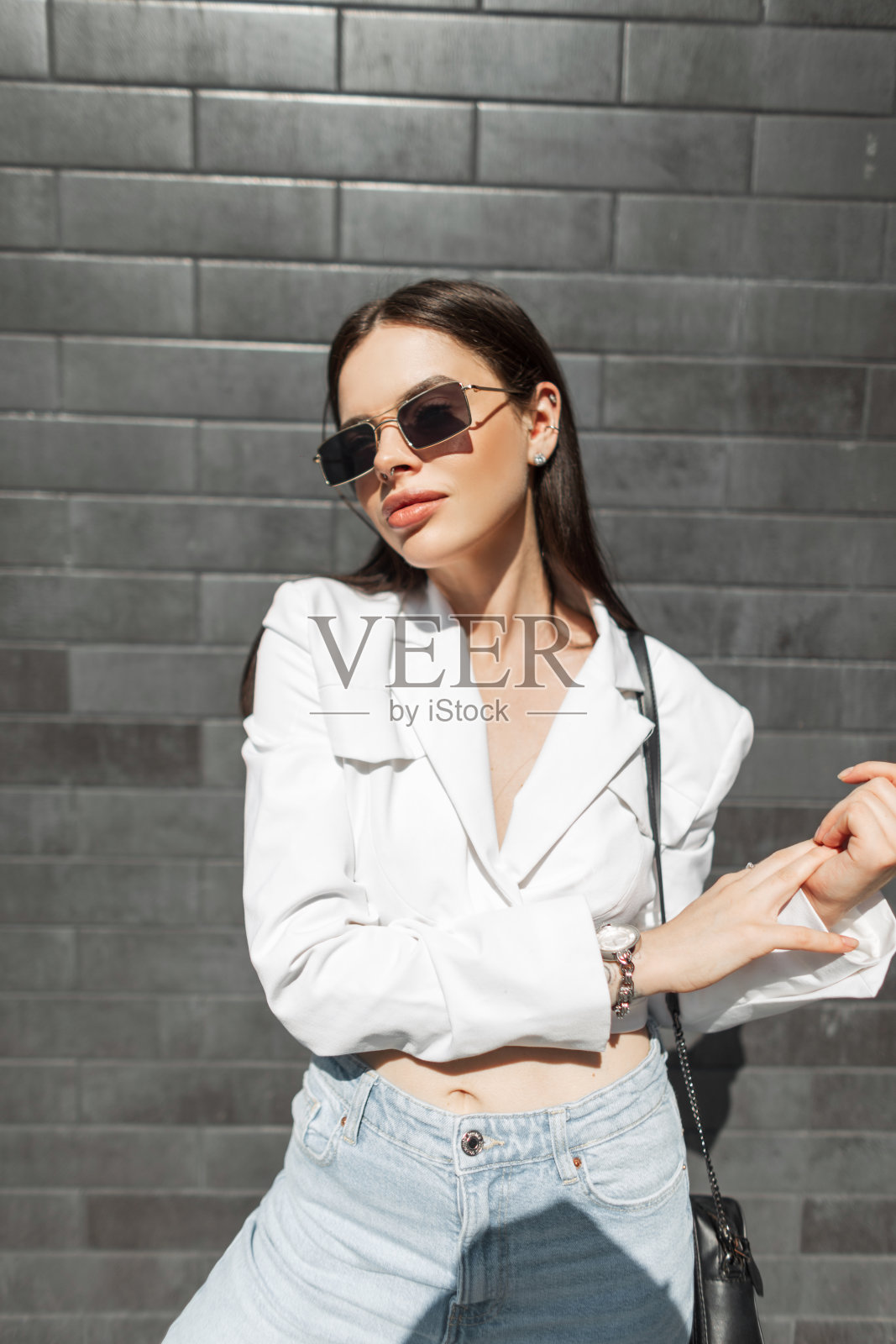 在一个阳光明媚的日子里，时尚美丽的城市商务女孩，戴着复古太阳镜，穿着白色上衣和牛仔裤，提着一个包，站在城市的砖墙旁照片摄影图片