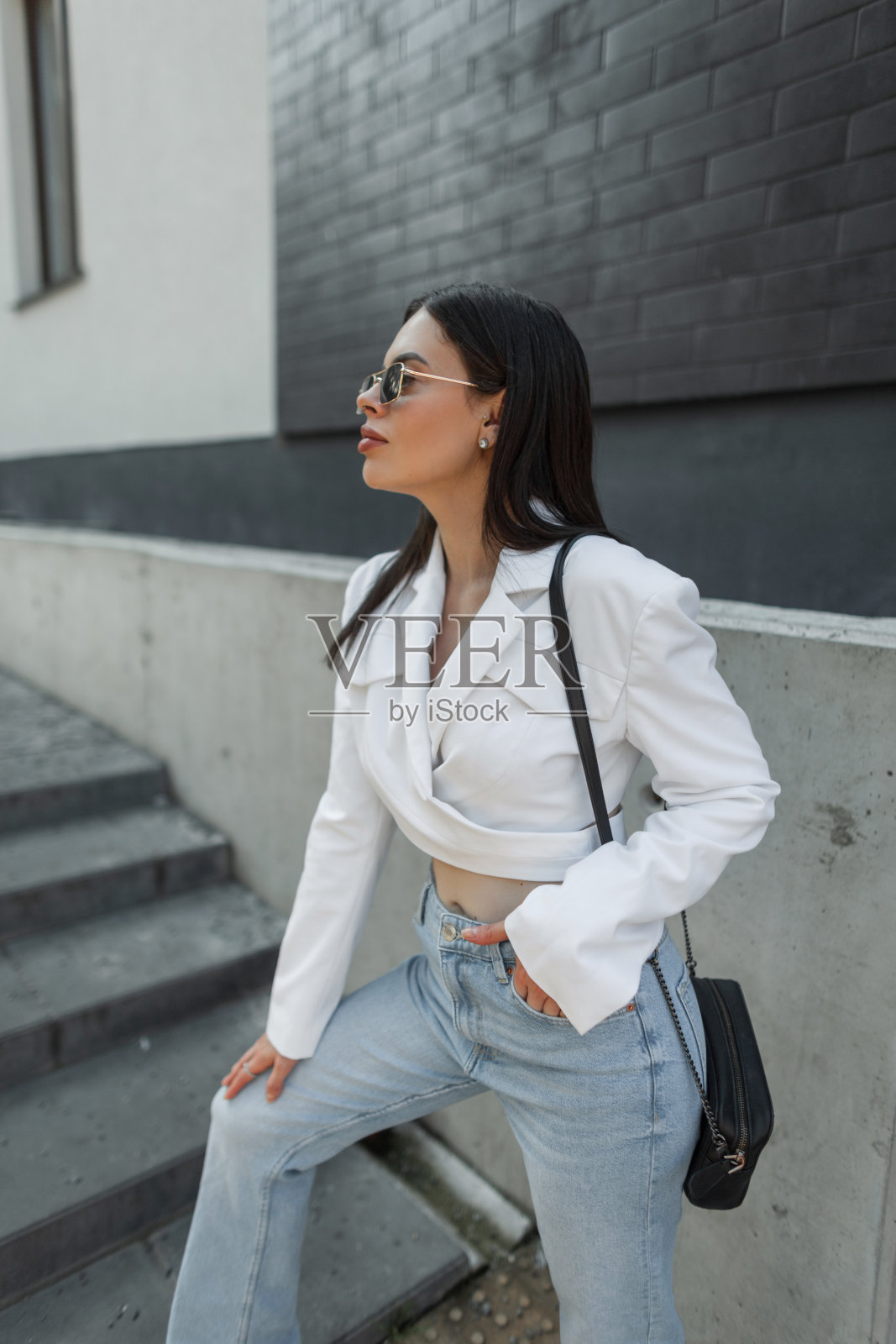 时尚美丽的年轻时尚的女孩戴着太阳镜，穿着时尚休闲的衣服，白色西装和复古的蓝色牛仔裤，带着一个包在城市附近的砖黑色的墙照片摄影图片