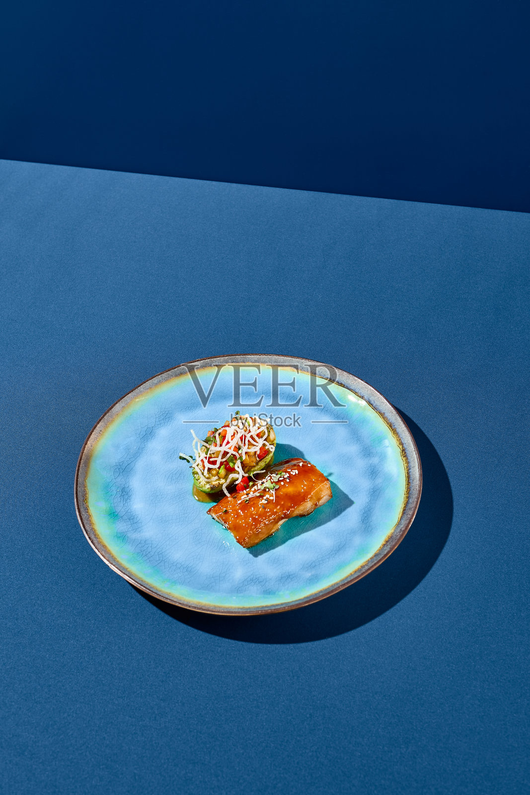 斜侧面垂直拍摄的鳕鱼在unagi酱伴随着烤鳄梨和蔬菜，优雅地呈现在一个蓝色的陶瓷盘子，极简主义风格的阴影在蓝色的背景照片摄影图片