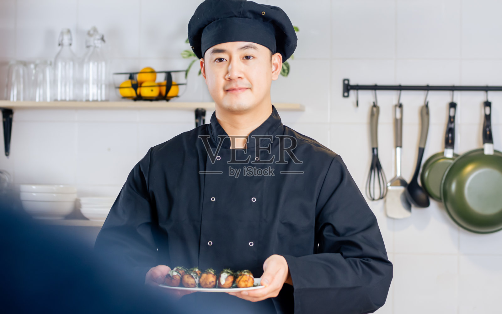 肖像中帅气的专业日本男厨师身穿黑色制服，头戴帽子，正在烹饪，展示着寿司街头小吃，微笑着自信、幸福地站在厨房里。餐厅，酒店概念。照片摄影图片