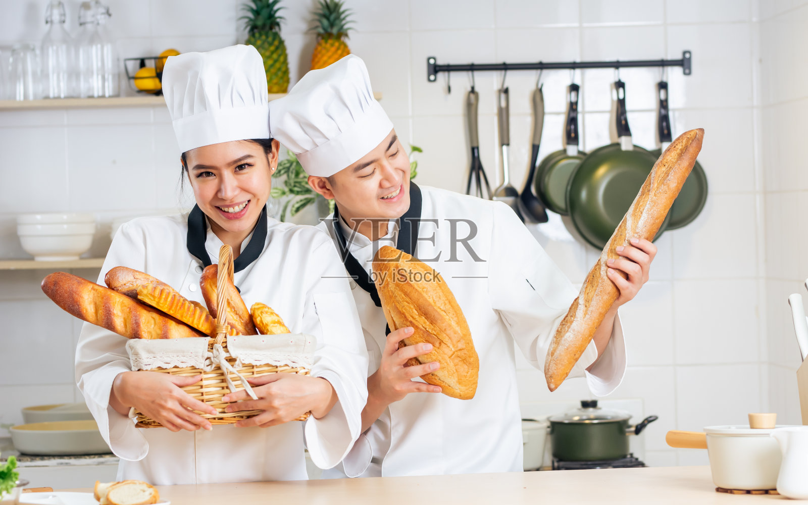 两位亚洲专业厨师身穿白色制服，头戴帽子，手拿法棍、面包，在厨房里开心地做着早餐。餐厅，酒店，食品概念。照片摄影图片