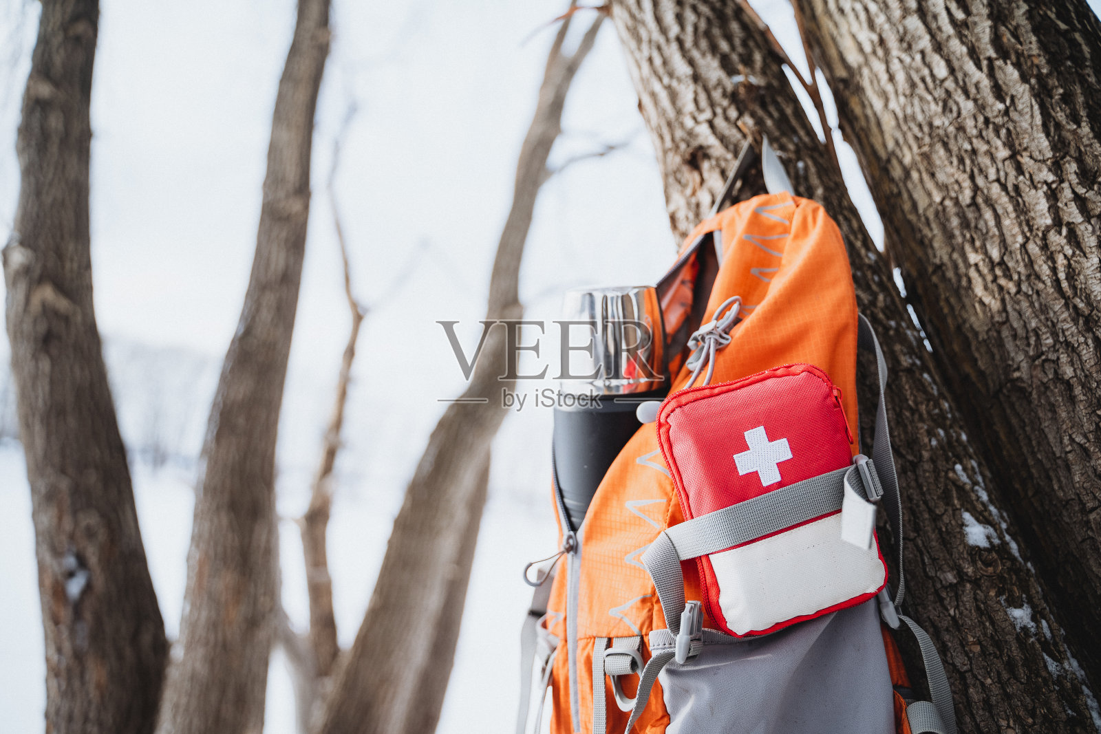在冬天的森林里，挂在树上的橙色背包里有一个急救箱和一个热水瓶。照片摄影图片