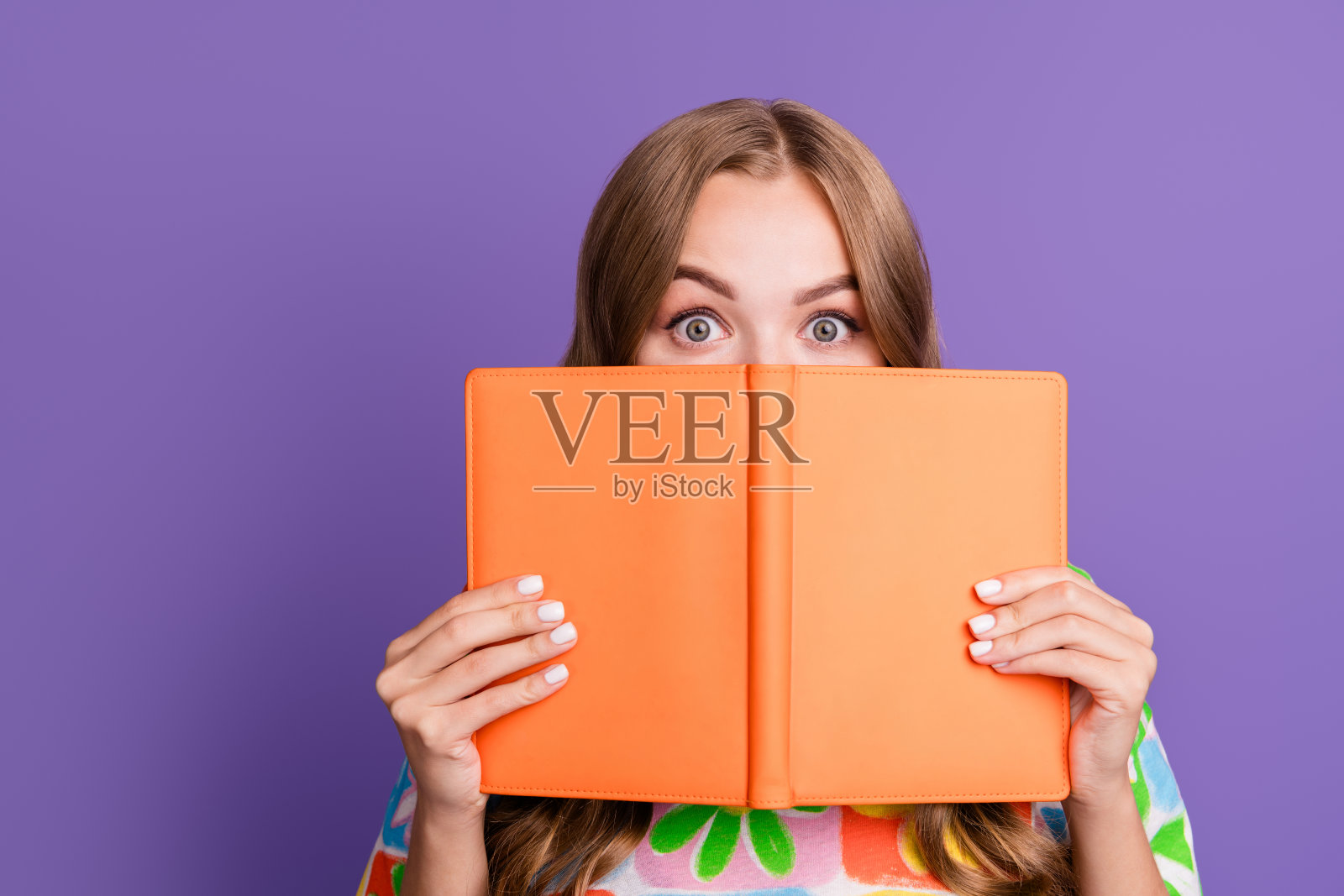 照片上的学生惊讶得说不出话来，一头卷发，穿着印花t恤，把脸藏在书后面，孤立在紫色的背景上照片摄影图片