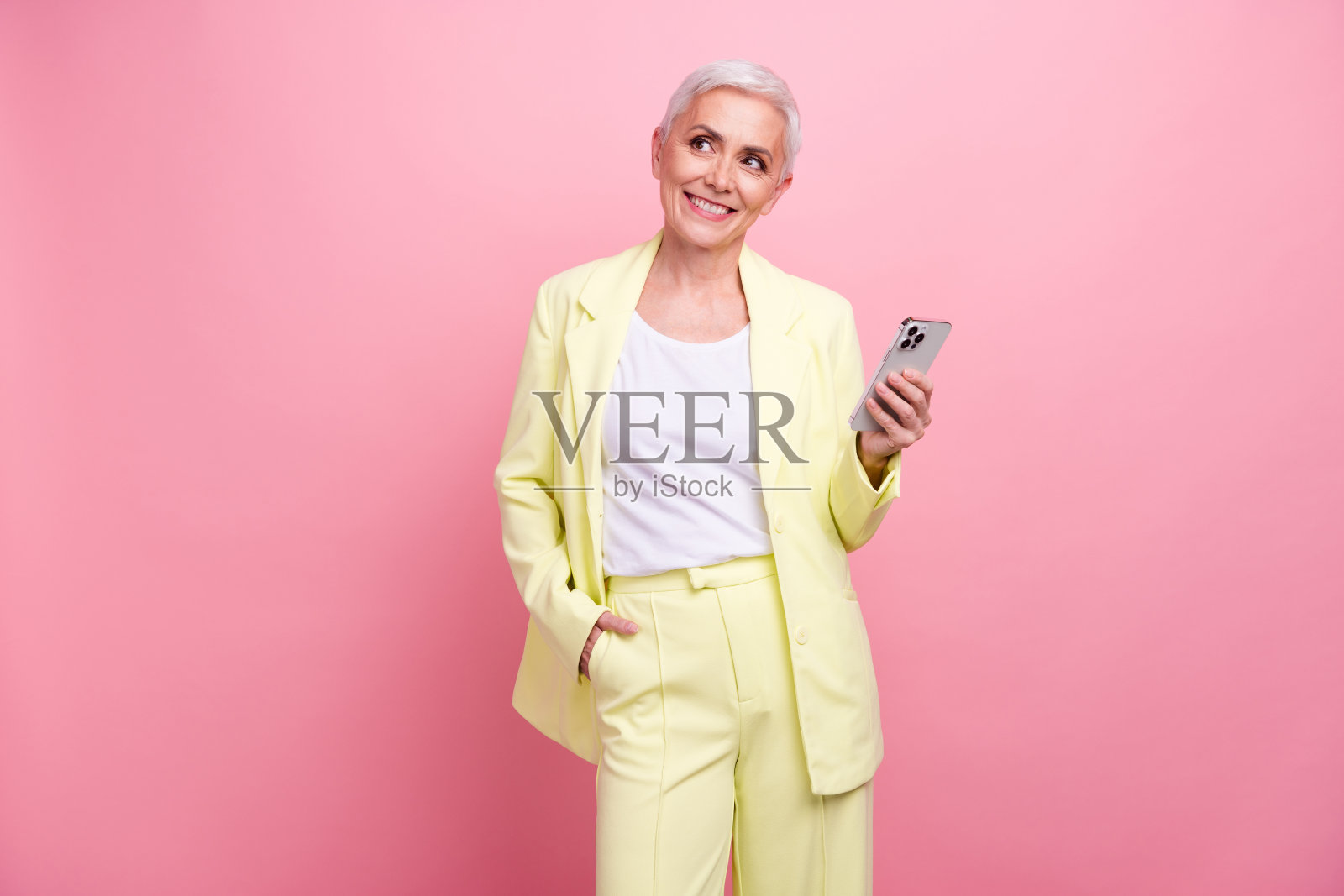照片中的老太太业务销售经理穿着黄色西装，用手机看起来空白的空间应用商店孤立在粉红色的背景照片摄影图片