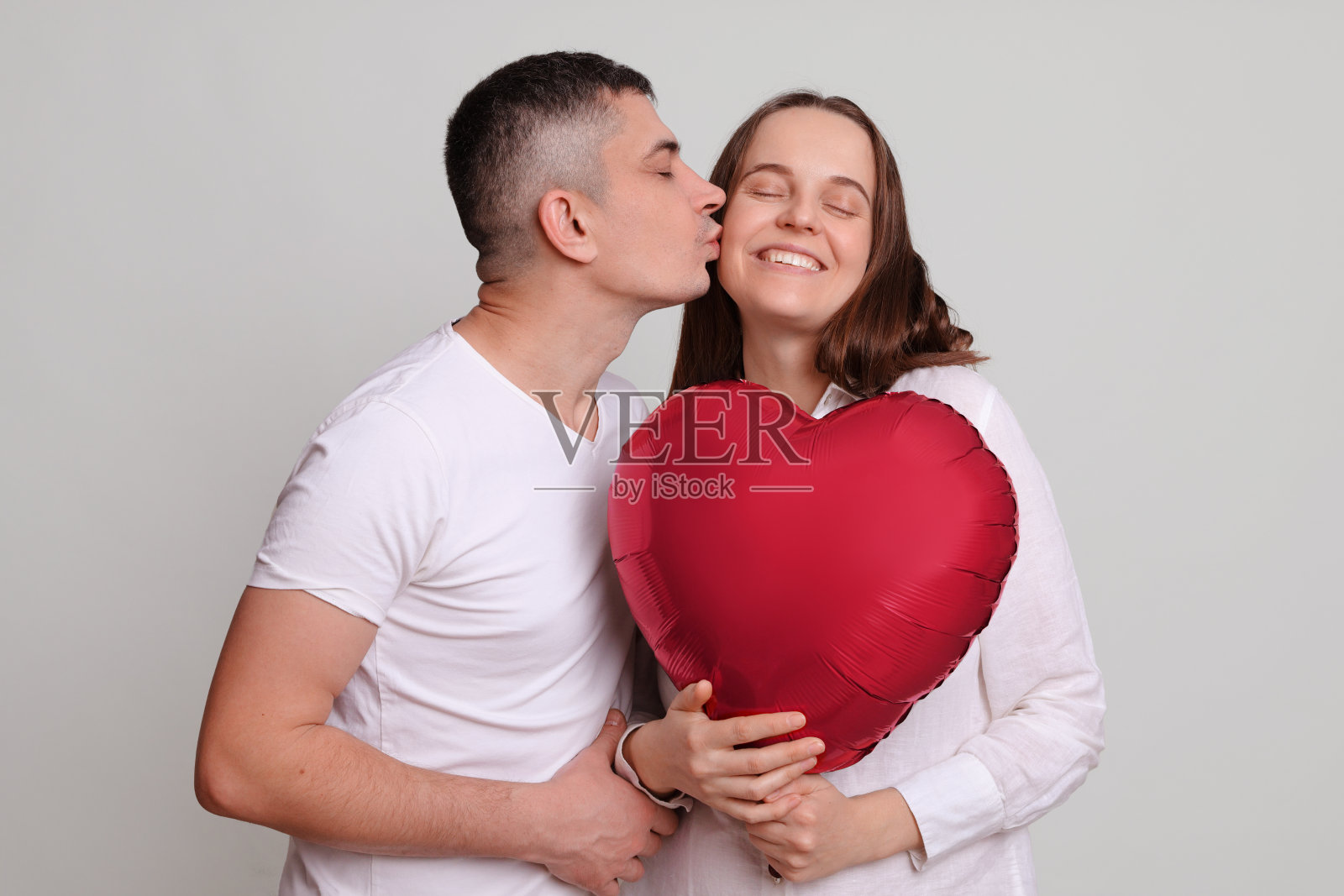 满意高兴的白人男人和女人抱着心形的气球孤立在灰色的背景英俊的男人亲吻心爱的女朋友脸颊祝贺节日照片摄影图片