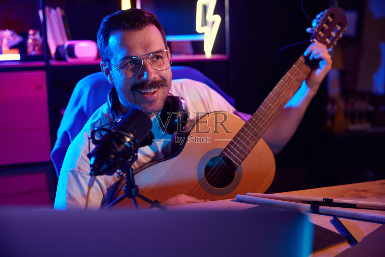 开朗的男人，吉他手在工作室的霓虹灯下用专业麦克风为他博客的追随者录制视频课程。照片摄影图片